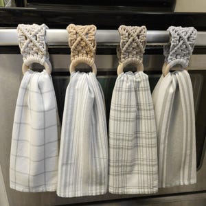 Pode incluir: Quatro panos de cozinha suspensos com topos em macram&eacute; nas cores creme, castanho, bege e cinzento claro. Cada pano apresenta um padr&atilde;o diferente, incluindo xadrez e riscas, preso a um anel de madeira natural num puxador de forno.