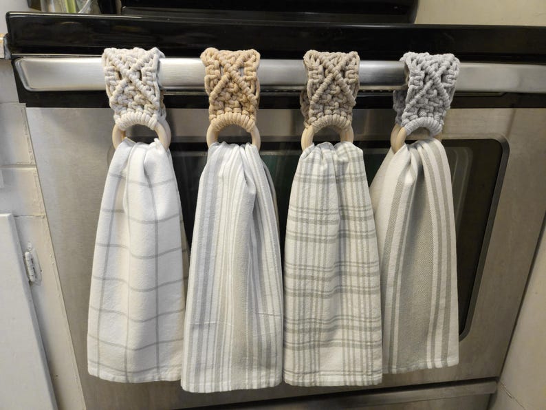Pode incluir: Quatro panos de cozinha suspensos com topos em macram&eacute; nas cores creme, castanho, bege e cinzento claro. Cada pano apresenta um padr&atilde;o diferente, incluindo xadrez e riscas, preso a um anel de madeira natural num puxador de forno.