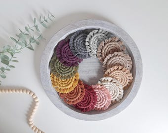 Pode incluir: Uma coleção de bases para copos circulares em macramé dispostas em círculo dentro de uma bandeja redonda de madeira cinza-claro. As bases apresentam uma variedade de cores, incluindo cinza-escuro, cinza-claro, bege, creme, rosa, vermelho, laranja, amarelo, oliva e roxo.