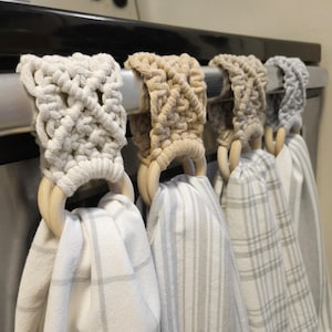 May include: Four macrame towel holders with wooden rings, suspended from a stainless steel bar. The macrame is in cream, beige, and grey tones. Each holder features a unique woven design. The towels are white with grey stripes and check patterns.