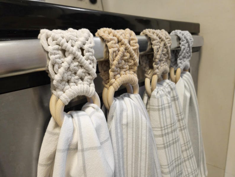 Pode incluir: Quatro panos de cozinha suspensos com topos em macram&eacute; em tons de creme, bege e cinzento. Cada pano apresenta um padr&atilde;o diferente, incluindo xadrez e riscas, fixado a um anel de madeira clara e lisa com um design de n&oacute;s de macram&eacute; decorativo.