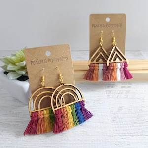 May include: Two pairs of dangle earrings on brown cardboard displays labeled Peach & Poppyseed. One pair features wooden rainbow arches with multi-colored fringe tassels. The other pair features wooden triangles with white, orange, and pink fringe.