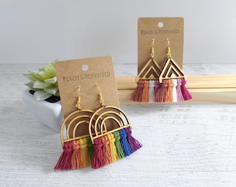 May include: Two pairs of dangle earrings on brown cardboard displays labeled Peach & Poppyseed. One pair features wooden rainbow arches with multi-colored fringe tassels. The other pair features wooden triangles with white, orange, and pink fringe.