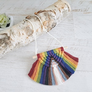 May include: A rainbow-coloured macrame wall hanging with wooden dowels and tassels. The hanging is made with a variety of colours, including red, orange, yellow, green, blue, indigo, and violet.