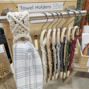Pode incluir: Uma cole&ccedil;&atilde;o de suportes para toalhas em macram&eacute; pendurados numa barra de metal prateado. Cada suporte apresenta um anel de madeira com um design de cord&atilde;o atado em v&aacute;rias cores, incluindo creme, cinzento, verde-azeitona e bordeaux. Um suporte exibe uma toalha branca aos quadrados.