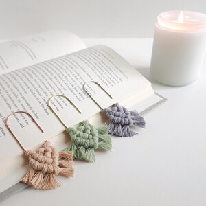 May include: Three metal bookmarks with macrame tassels in beige, sage green, and slate blue. Each bookmark features a U-shaped wire frame with a knotted cotton cord design attached to the bottom, resting on the pages of an open hardcover book.