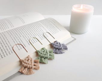 Pode incluir: Três marcadores de página de metal em forma de clipe com borlas decorativas de macramé em tons suaves de bege, verde sálvia e azul acinzentado. Cada marcador está preso a um design de cordão de algodão com nós na página de um livro.