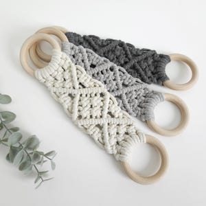 Pode incluir: Tr&ecirc;s tiras de macram&eacute; com padr&otilde;es de n&oacute;s intrincados em creme, cinzento claro e cinzento carv&atilde;o escuro. Cada tira est&aacute; presa a dois an&eacute;is de madeira clara, lisos e circulares, em ambas as extremidades, criando um design de la&ccedil;o funcional.