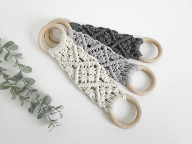 Pode incluir: Tr&ecirc;s tiras de macram&eacute; com padr&otilde;es de n&oacute;s intrincados em creme, cinzento claro e cinzento carv&atilde;o escuro. Cada tira est&aacute; presa a dois an&eacute;is de madeira clara, lisos e circulares, em ambas as extremidades, criando um design de la&ccedil;o funcional.