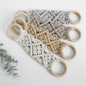 Pode incluir: Tr&ecirc;s al&ccedil;as de macram&eacute; com n&oacute;s complexos em forma de diamante em cord&atilde;o de algod&atilde;o nas cores creme, bege e cinza. Cada al&ccedil;a est&aacute; presa a dois an&eacute;is de madeira circulares e lisos em ambas as extremidades, criando um design funcional para pendurar.