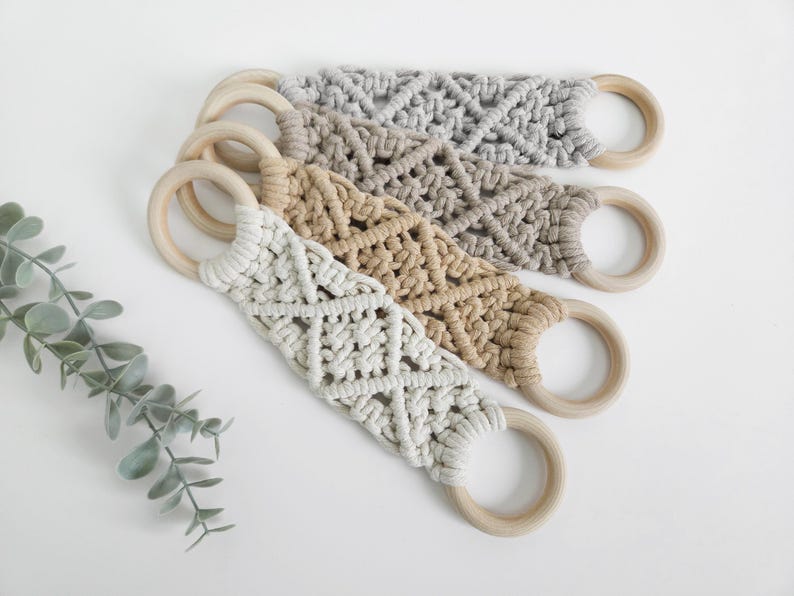 Pode incluir: Tr&ecirc;s al&ccedil;as de macram&eacute; com n&oacute;s complexos em forma de diamante em cord&atilde;o de algod&atilde;o nas cores creme, bege e cinza. Cada al&ccedil;a est&aacute; presa a dois an&eacute;is de madeira circulares e lisos em ambas as extremidades, criando um design funcional para pendurar.