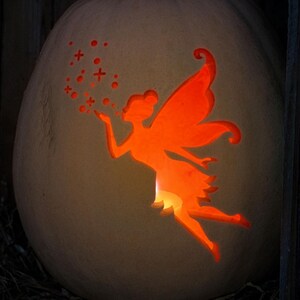 May include: A carved pumpkin with a fairy design lit from inside. The fairy is blowing stars, and the pumpkin rests on hay. The pumpkin is displayed in a dark wooden structure, creating a Halloween theme.