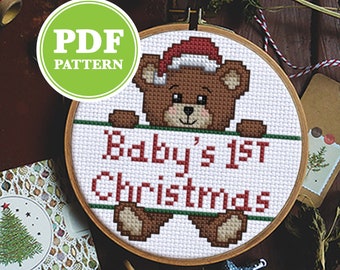 May include: A cross-stitch pattern for a baby's first Christmas. The design features a brown teddy bear wearing a Santa hat, holding a banner with the text "Baby's 1st Christmas". The pattern is in a wooden embroidery hoop, with other craft supplies in the background.