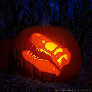Könnte beinhalten: Ein großer, orangefarbener Kürbis mit einem geschnitzten Dinosaurierschädel-Design. Das Innere des geschnitzten Schädels leuchtet mit einem warmen Licht und schafft eine gruselige Halloween-Dekoration. Der Hintergrund ist dunkel.