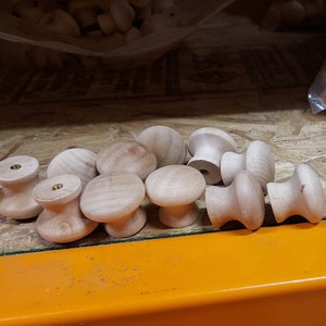 Puede incluir: Primer plano de un montón de pomos de madera, algunos con tornillos de latón, sobre una superficie de madera. Los pomos son de color marrón claro y tienen un acabado liso. La superficie es de color marrón claro y tiene una textura rugosa. Los pomos están dispuestos en un patrón aleatorio. El texto "UN1" es visible en una etiqueta en la superficie naranja.