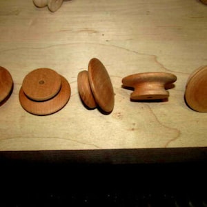 May include: A collection of unfinished wooden knobs and pulls in various shapes, including round, stacked, and mushroom-shaped designs. The natural wood grain is visible, and the pieces are arranged on a light-colored wooden surface.