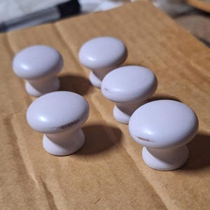 May include: Six white ceramic knobs with a smooth, rounded surface. The knobs are arranged in a semi-circle on a light brown surface.