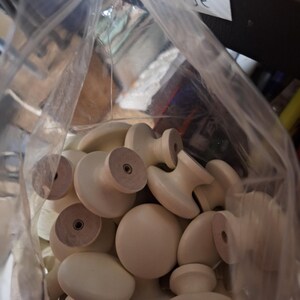 May include: A clear plastic bag filled with numerous off-white wooden knobs. The knobs have various shapes, including rounded and mushroom-style designs. The bag is partially transparent, and a handwritten label is visible.