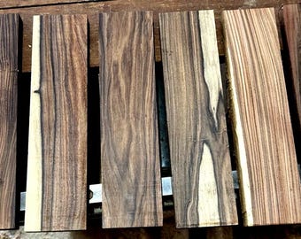 Könnte beinhalten: Sechs rechteckige Holzbretter in Braun- und Cremetönen. Die Bretter weisen unterschiedliche Maserungsmuster auf, einige mit dunklen, fast schwarzen Streifen. Das Holz scheint unbehandelt zu sein.