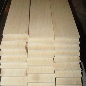 May include: Stacks of unfinished, light-colored wooden planks. The planks are rectangular and appear to be made of softwood. The wood grain is visible, and the planks are stacked neatly, ready for use in a woodworking project.