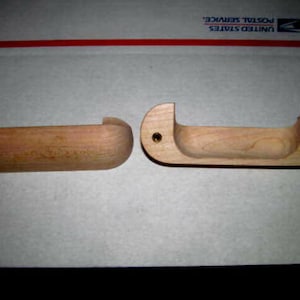 May include: Two unfinished wooden drawer pulls. One is a solid, rounded shape, while the other has a recessed handle area and two brass-colored screw holes. Both are made of light-colored wood.