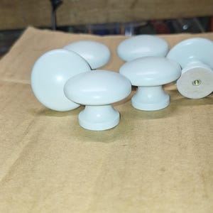 May include: A set of seven white ceramic cabinet knobs with a rounded top and a small base. The knobs are arranged on a brown surface.