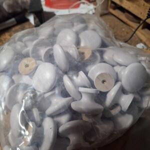 May include: A plastic bag filled with a large quantity of white ceramic cabinet knobs. The knobs have a simple, round design with a small wooden base.
