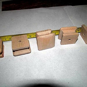 May include: Five small, unfinished wooden blocks of varying designs. Each block is a natural wood color and has a hole drilled through it. A yellow measuring tape is partially visible in the background.