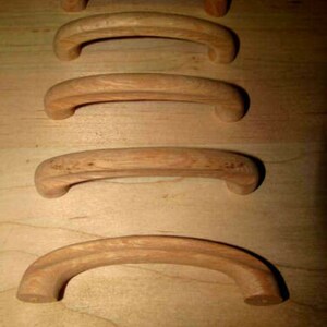 May include: Five curved wooden cabinet handles. The handles are a light brown color and have a smooth, polished finish. The handles are arranged in a vertical row, showcasing their arched design. These handles are suitable for furniture or cabinet projects.