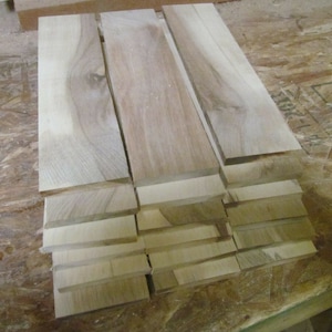 May include: A collection of unfinished wooden planks, varying in size, stacked and arranged on a light-colored surface. The wood has a natural, light brown color with visible grain patterns, suggesting it is ready for woodworking projects.