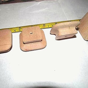 May include: Four unfinished wooden cabinet knobs in various shapes. The knobs are light brown and appear to be made of a soft wood. A yellow measuring tape is in the background, showing inches.