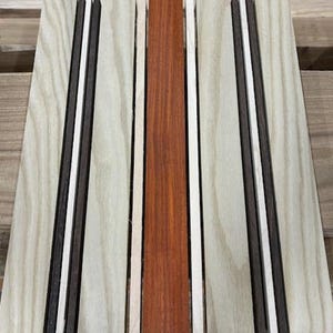May include: Close-up of a wooden board with five strips of wood. The strips are different colors and widths. The strips are arranged in a vertical pattern. The wood is light brown, dark brown, white, and red.