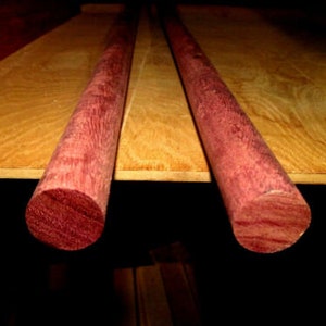 May include: Two round wooden dowels with a reddish-purple hue and a wood grain pattern. The dowels are lying on a light brown surface.