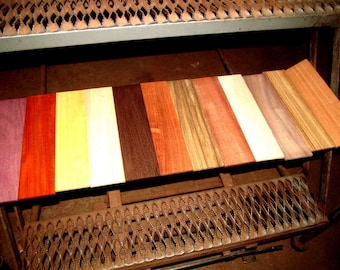 May include: A collection of various wood planks, each displaying a unique color and grain pattern. The planks range from deep purple to light yellow and brown, showcasing the natural diversity of wood. The planks are arranged on a metal surface.