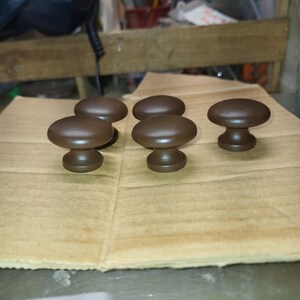 May include: Five brown wooden cabinet knobs with a rounded top and a small base. The knobs are arranged in a semi-circle on a brown paper surface.