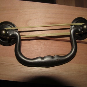 May include: Close-up of a dark metal drawer pull with a curved handle and circular mounting plates. A light-colored rubber band is stretched across the handle. The drawer pull is mounted on a light brown wooden surface.
