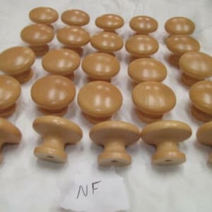 Puede incluir: Un conjunto de veinticuatro pomos de madera marrón claro. Cada pomo tiene una parte superior redondeada y una base cilíndrica corta. La veta de la madera es visible. Los pomos están dispuestos sobre una superficie blanca.