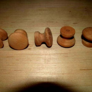 May include: Five wooden cabinet knobs in various designs. The knobs are made of light brown wood and are arranged in a row. The knobs have different shapes, including round, mushroom, and hourglass styles. These knobs are suitable for furniture restoration or DIY projects.