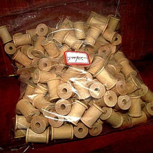 May include: A clear plastic bag filled with numerous small, light brown wooden spools. Each spool has a cylindrical body with a central hole and a slightly flared base. A small red and white tag is visible.