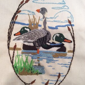 May include: Embroidered artwork depicting three ducks in a pond. The ducks are gray, green, and white with brown and blue accents. The pond is blue with brown reeds and green grass on the shore.