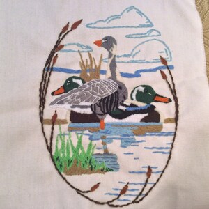 May include: Embroidered wall art featuring three ducks in a pond setting. The ducks are a grey goose and two mallards. The background is a white fabric with blue and brown embroidery.