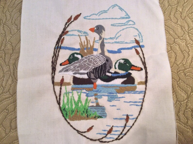 May include: Embroidered wall art featuring three ducks in a pond setting. The ducks are a grey goose and two mallards. The background is a white fabric with blue and brown embroidery.