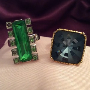 May include: Two rings with large, colorful gemstones. The first ring has a rectangular green stone surrounded by smaller silver stones. The second ring has a square, dark blue stone with a faceted design, surrounded by a gold band.
