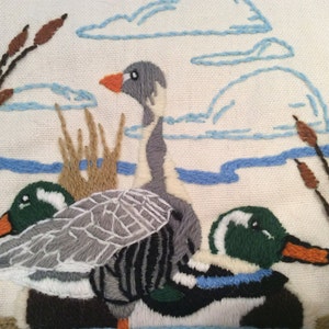 May include: Embroidered wall hanging featuring three geese in a pond setting. The geese are gray, green, and white with brown and blue accents. The background is a cream-colored fabric with blue embroidered clouds and brown reeds.