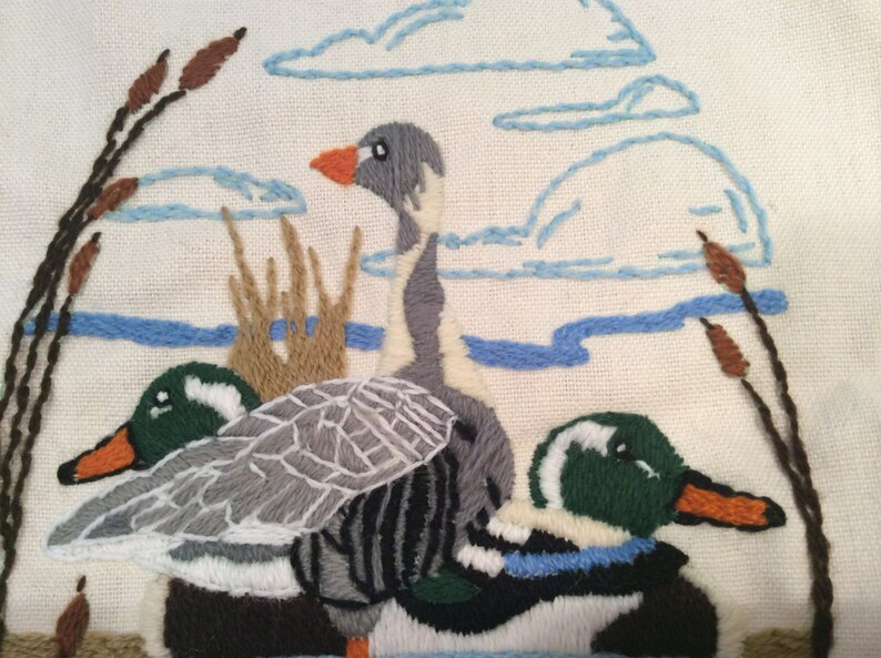 May include: Embroidered wall hanging featuring three geese in a pond setting. The geese are gray, green, and white with brown and blue accents. The background is a cream-colored fabric with blue embroidered clouds and brown reeds.