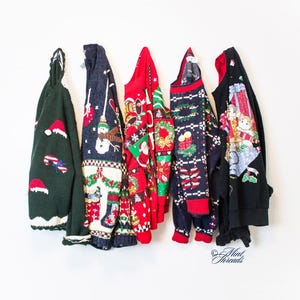 May include: Five Christmas sweaters hang against a white wall. The sweaters are various colors, including green, red, and navy, and feature holiday-themed designs like snowmen, Santa, and candy canes. The sweaters are made of a knitted material.