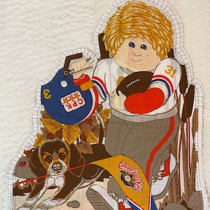 Pode incluir: Painel de tecido com uma ilustração de desenho animado de um menino com uniforme de futebol segurando uma bola de futebol, com um cachorro na coleira. O menino usa um capacete e o painel tem o texto "Cabbage Patch Kids" em uma flâmula. As bordas são inacabadas.