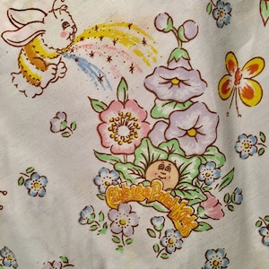 Könnte beinhalten: Weißer Stoff mit einem sich wiederholenden Muster aus Zeichentrickfiguren, Blumen und Schmetterlingen. Das Design umfasst ein Häschen, das einen Regenbogen bläst, ein Cabbage Patch Kids-Logo und florale Elemente in Pastellfarben.