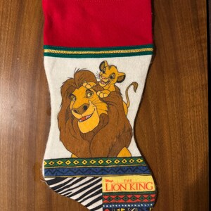 May include: A Christmas stocking featuring a design from Disney's The Lion King. The stocking has a red cuff, a white body with Simba and Mufasa, and a patterned bottom with the movie's logo. A gold and green trim separates the cuff from the body.