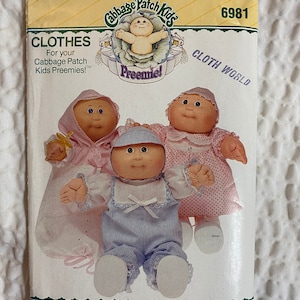 Könnte beinhalten: Ein Vintage-Nähmuster für Cabbage Patch Kids zum Nähen von Kleidung für Frühchenpuppen. Das Muster ist für ein blau-weiß gestreiftes Outfit, ein rosa-weiß gepunktetes Kleid und ein rosa-weißes Kleid mit einer gelben Schleife. Das Muster stammt aus der Cloth World-Kollektion und trägt die Nummer 6981.